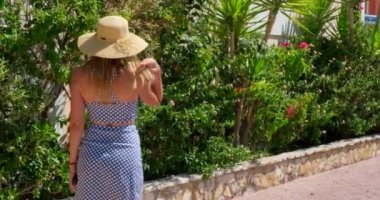 Elbise ve hasır şapka giymiş bir kadın bahçede yürüyor. Yaz zamanı. Kadın otelin bölgesine giriyor. Ağır çekim. Kız kameraya bakıyor.