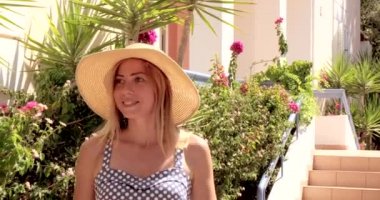 Elbise ve hasır şapka giymiş bir kadın bahçede yürüyor. Yaz zamanı. Kadın otelin bölgesine giriyor. Ağır çekim. Kız kameraya bakıyor.