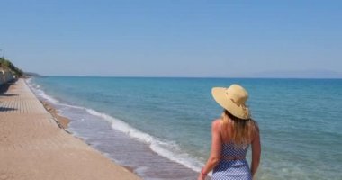 Elbise giymiş genç bir kadın yaz plajında yürüyor. Hasır şapkalı kız denizin kenarında yürüyor.