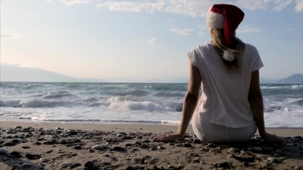 戴着圣诞老人帽子的女人坐在沙滩上的沙子上. 穿着白衣的女孩坐在大海的背景上。 热带圣诞假期的概念。 在阳光充足的热带海滨度假胜地度假 — 图库视频影像
