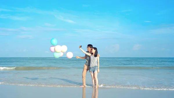 Feliz casal asiático segurando balões coloridos na praia durante — Fotografia de Stock