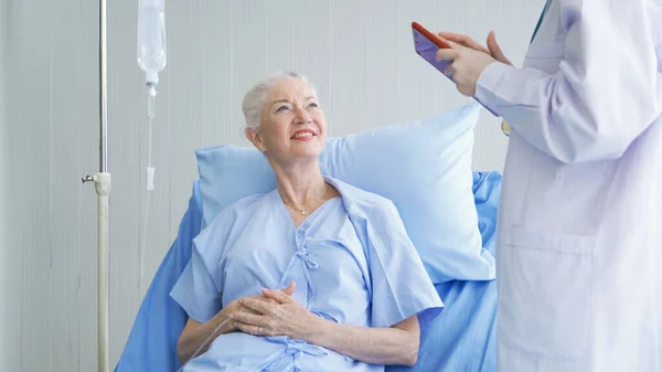 Happy woman doctor talking and using tablet to sick old female senior elderly patient lying in bed in hospital ward room in medical, technology and healthcare treatment concept. Caucasian people.