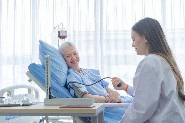Happy woman doctor checking blood pressure test of sick old female senior elderly patient lying in bed in hospital ward room in medical, and healthcare treatment concept. Caucasian white people.