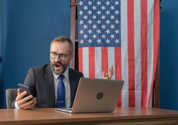 Business white vintage american man, caucasian person looking at mobile phone, thinking about problem with computer notebook laptop and suffering from depression with american or usa national flag.