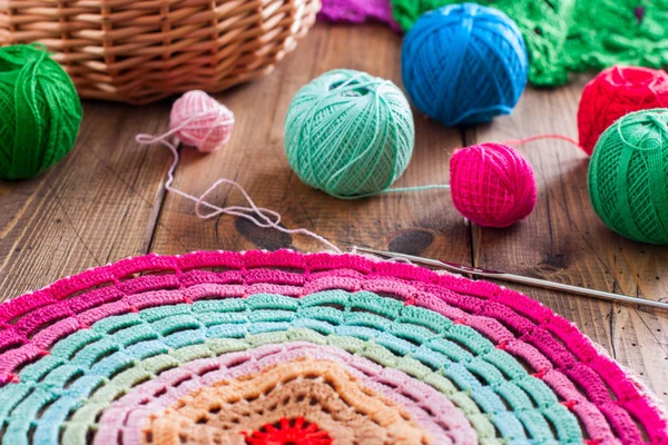 Multi-colored balls of cotton thread for crocheting in a wicker basket on a wooden table and ready knitted napkin made of multi-colored thread, selective focus