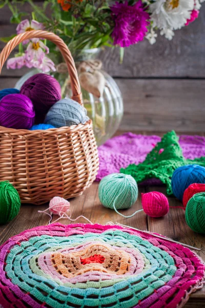 Multi-colored balls of cotton thread for crocheting in a wicker basket on a wooden table and ready knitted napkin made of multi-colored thread, selective focus