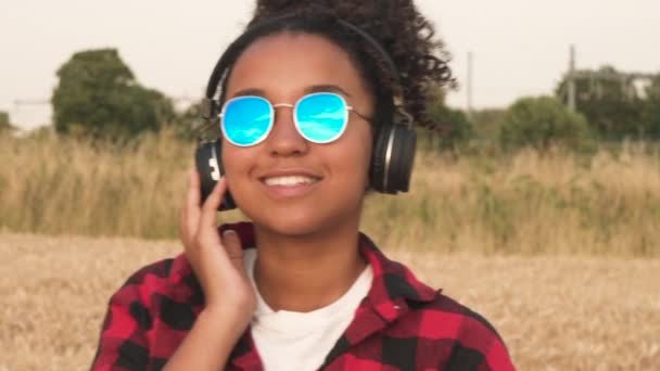 慢动作视频美丽的混合种族非洲裔美国女孩十几岁的年轻女子穿着红色和黑色的衬衫和蓝色的太阳镜听音乐在她的手机和无线耳机 — 图库视频影像