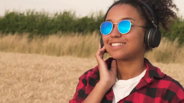 赤と黒のシャツと彼女の携帯電話と無線のヘッドフォンで音楽を聞いて青いサングラスを身に着けている美しい混血アフリカ系アメリカ人の女の子 代若い女性のスロー モーション ビデオ — ストック動画