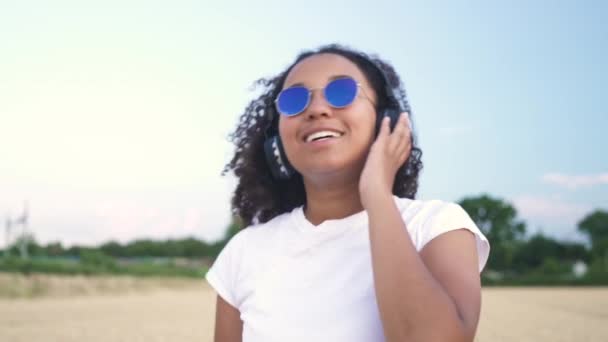 シャツと青いサングラス無線ヘッドフォンで音楽を聴き歩いて美しい混血アフリカ系アメリカ人の女の子 代若い女性のスローモーション フォロー ショット ビデオ — ストック動画