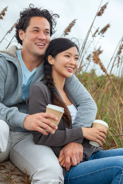 アジア中国男と女 少年少女カップル完璧な歯で笑顔で飲む紅茶やコーヒーのテイクアウト カップのビーチに座っています — ストック写真