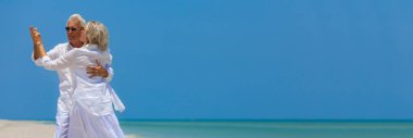 Panoramic banner image of happy senior man and woman couple dancing and holding hands on a deserted tropical beach with turquoise sea and clear blue sky clipart