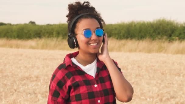 スローモーション美しい混血アフリカ系アメリカ人女の子ティーンエイ ジャー若い女性青いサングラスをかけて夕日や日の出無線ヘッドフォンで音楽を聴き歩いてのビデオ クリップを追跡 — ストック動画
