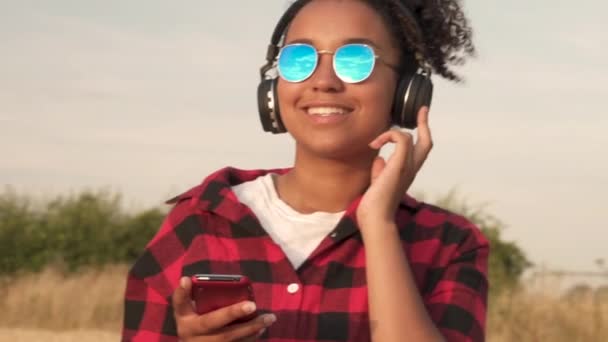 赤と黒のシャツと彼女の携帯電話と無線のヘッドフォンで音楽を聞いて青いサングラスを身に着けている美しい混血アフリカ系アメリカ人の女の子 代若い女性のスロー モーション ビデオ — ストック動画