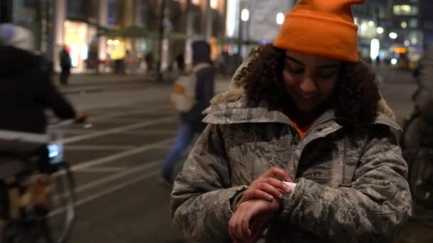 美丽的双种族非洲裔美国少女少女在城市街头在冬天的夜晚戴着橙色帽子和迷彩夹克使用她的智能手表或智能手表 — 图库视频影像