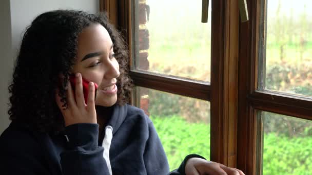 青いパーカーを着て 彼女の携帯電話で話している窓のそばに座って 幸せな笑顔美しい混合レースアフリカ系アメリカ人の女の子の若い女性 — ストック動画