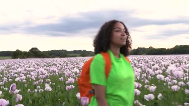 美しい幸せな混合レースアフリカ系アメリカ人の女の子の女の子の若い女性は ピンクのケシの花のフィールドを通ってパス上のオレンジ色のバックパックでハイキング — ストック動画
