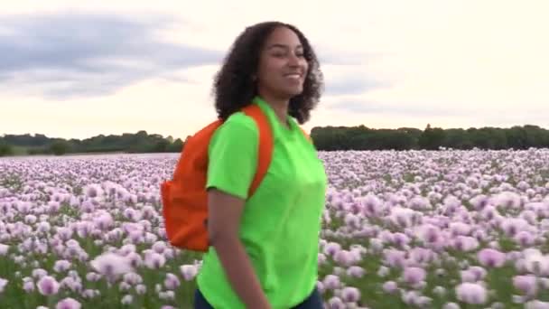 美丽的快乐混合种族非洲裔美国少女少女女青年徒步旅行与橙色背包通过粉红色罂粟花领域路径 — 图库视频影像