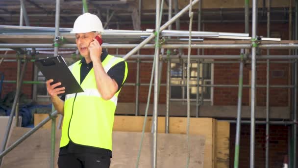 男性ビルダーの責任者 労働者 測量士 エンジニア または建築家は クリップボードを保持し 携帯電話で話す足場に立って建設建設現場に取り組んでいます — ストック動画