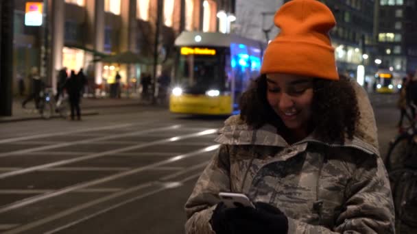 美丽的混合种族女少女年轻女子穿着迷彩服 橙色的贝尼帽 晚上使用她的手机测试消息或社交媒体与电车亚历山大广场站 — 图库视频影像