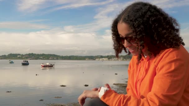漂亮的混血非洲裔美国少女 身穿橙色帽衫 坐在海滨 用她的智能手表进行社交媒体 交流或发短信 — 图库视频影像