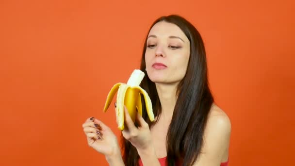 Mujer joven y saludable con piel perfecta comiendo plátano sobre fondo naranja brillante. Frutas tropicales. Concepto de Comida Vegetariana — Vídeos de Stock