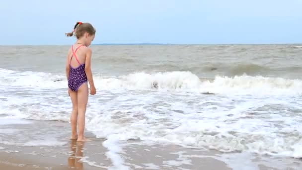 4K. Une petite fille en maillot de bain joue avec les vagues sur le rivage de l'océan. En mer, une tempête faible, un vent fort et des vagues. Vacances d'été, vacances en famille — Video