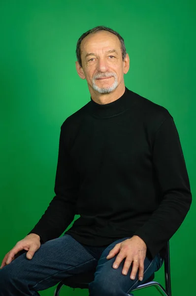 Портрет пожилого седого человека с хорошо отделанной бородой на зеленом фоне в черном свитере и сидящего на стуле в студии — стоковое фото