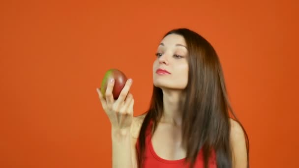 Rapariga atraente com cabelo escuro comprido segurando amarelo-vermelho manga na mão e tentando comer esta fruta tropical em fundo laranja no estúdio — Vídeo de Stock