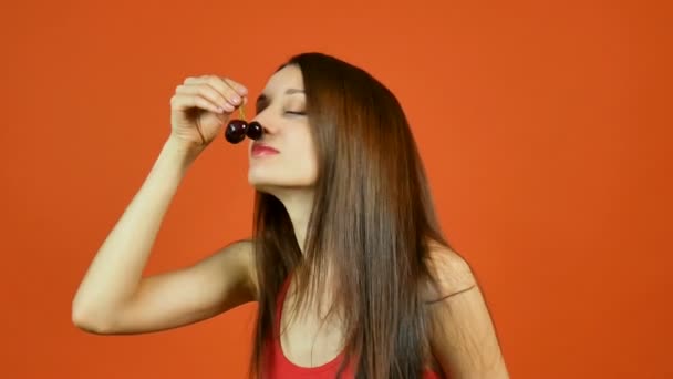 Ritratto di giovane donna sorridente che mangia due ciliegie rosso scuro su sfondo arancione in studio. Alimentazione sana . — Video Stock