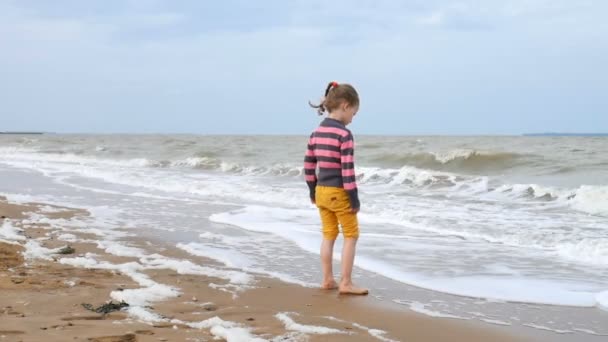 Lustiges Mädchen in Shart und gelber Hose, das die Wellen im Ozean betrachtet, die am Wasser stehen. ein schwacher Sturm auf See, starker Wind und Wellen. — Stockvideo