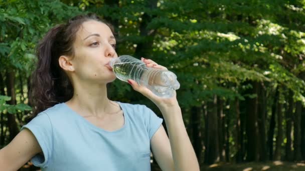 Fitness bruneta žena pitnou vodu venku v parku po její spuštění na pozadí zelených stromů. — Stock video