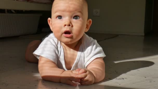 Carino bambino di sei mesi è sdraiato sul pavimento sorridendo allegramente. Il bambino bussa un ruscello sul pavimento e strattona vigorosamente le gambe — Video Stock