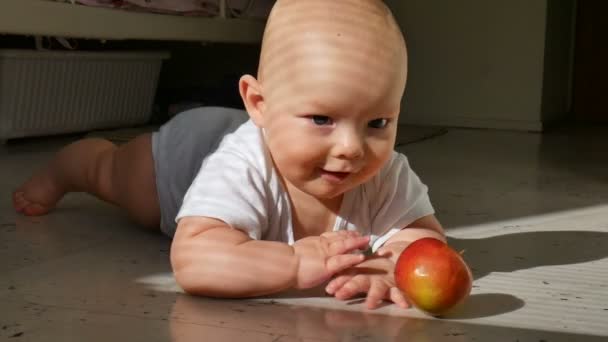 Il bambino è sdraiato sul pavimento e tiene una mela rossa. Il bambino è molto vivace e allegro, bussa attivamente con mani e piedi sul pavimento. — Video Stock
