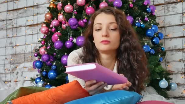 Hermosa mujer sexy con árbol de Navidad en el fondo leyendo un libro acostado sobre almohadas brillantes. Retrato de una chica posando bonita. Atractiva morena femenina relajante — Vídeo de stock