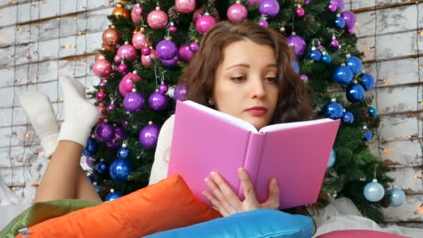 Jovem morena está lendo um livro no fundo de uma árvore de Natal elegante. Cores não convencionais, decorações criativas para o Natal — Vídeo de Stock