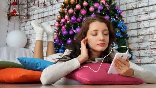 Привлекательная брюнетка лежит на ярких подушках возле елки и слушает музыку. Необычные рождественские украшения, Вайолет, Новый год. 4K — стоковое видео