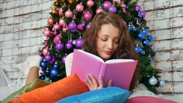 若いブルネットは、エレガントなクリスマス ツリーの背景に本を読んでいます。独創的な色、クリスマスのための創造的な装飾 — ストック動画