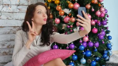 Noel kavramı - genç kadın dekore edilmiş Noel ağacı yakınındaki selfie fotoğraf çekiyorum. Kıvırcık kumral bir zafer jest yapma