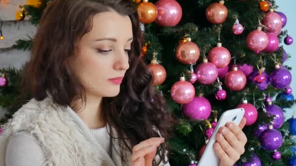 Красивая брюнетка на фоне украшенной елки, листая меню в своем смартфоне. проверяет почту, новогодние поздравления, интернет-магазины — стоковое видео