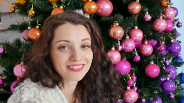 Weihnachten Konzept, schöne lockige Brünette lächelnd auf dem Hintergrund eines kreativ geschmückten Weihnachtsbaums. Porträt aus nächster Nähe — Stockvideo
