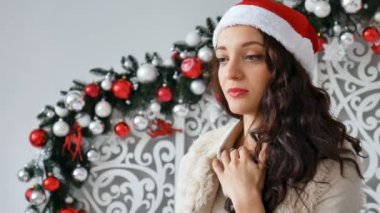 Üzgün Santas şapkalı güzel esmer düşünceli Noel dekorasyonları zemin karşı ayakta onun önünde görünüyor.