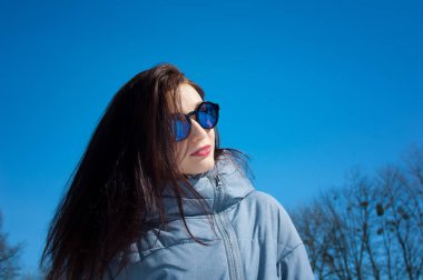 Uzun saçlı esmer yansıtılmış mavi güneş gözlüğü ve parlak mavi gökyüzü karşı kış giysileri portresi kapatın. Kış tatili, bahar için bekliyor.