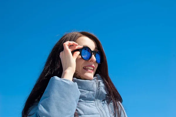 Retrato próximo de uma morena de cabelos longos em óculos de sol azuis espelhados e roupas de inverno contra um céu azul brilhante. Férias de inverno, esperando pela primavera — Fotografia de Stock