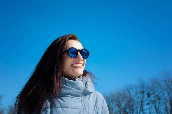 Utomhus livsstil närbild porträtt av vacker flicka vandrar i den snöiga vinter parken. Ler och njuter av vintern. Klädd i snygg speglad solglasögon, blå trenchcoat — Stockfoto