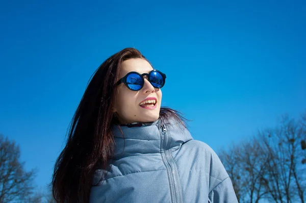 Retrato próximo de uma morena de cabelos longos em óculos de sol azuis espelhados e roupas de inverno contra um céu azul brilhante. Férias de inverno, esperando pela primavera — Fotografia de Stock