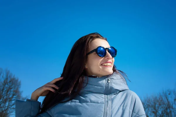 Близкий портрет длинноволосой брюнетки в зеркальных синих солнцезащитных очках и зимней одежде на фоне ярко-синего неба. Зимние каникулы, в ожидании весны — стоковое фото