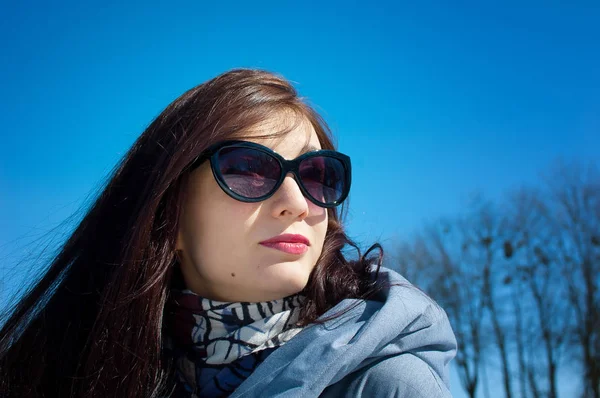 Uzun saçlı esmer yansıtılmış mavi güneş gözlüğü ve parlak mavi gökyüzü karşı kış giysileri portresi kapatın. Kış tatili, bahar için bekliyor