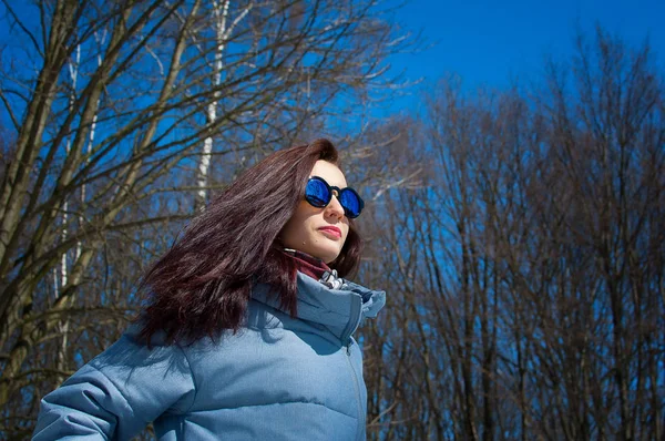 Retrato de una morena de pelo largo con gafas de sol azules espejadas y ropa de invierno contra un cielo azul brillante y árboles desnudos. Vacaciones de invierno, esperando la primavera — Foto de Stock