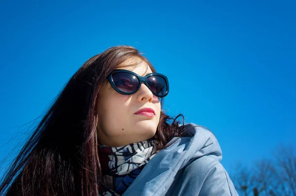Cute young gilr wearing dark sunglasses posing outdoors over blue sky during early spring time. — Stock Photo, Image