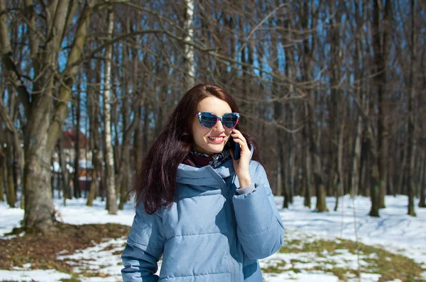 Leende ung kvinna i fashionabla solglasögon och vinterkläder talar via telefon medan spendera tid utomhus i parken under tidigt våren — Stockfoto